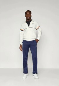 Λευκό φούτερ με φερμουάρ, με ναυτικές και πορτοκαλί ρίγες, συνδυασμένο με ναυτικά παντελόνια και λευκά αθλητικά παπούτσια. Διαθέτει λογότυπο στο φούτερ και στα παντελόνια.