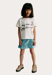 Vit T-shirt med kort ärm och svart broderad text samt rosett-design, kombinerad med en ljusblå kjol med volang nederkant och beige skor.