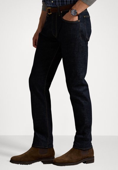 Jeans in denim blu scuro con cuciture arancioni, taglio slim, abbinati a stivali alla caviglia in suede marrone. Presenta tasche anteriori e una cintura marrone.