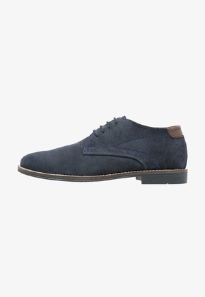 Chaussure oxford en daim marine avec une forme élégante, accents en cuir marron, cinq œillets et une semelle marron contrastante. Surface texturée.