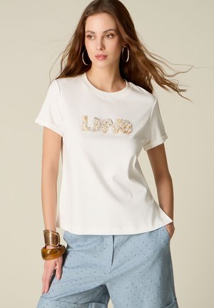 Młoda kobieta z długimi włosami, ubrana w białą koszulkę z cekinowym napisem „Love” oraz niebieskie spodnie w kropki, uzupełniona dużymi bransoletkami i kolczykami-kołami.