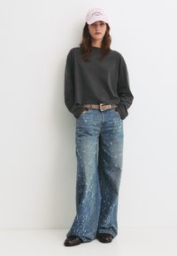 Maglietta grigia a maniche lunghe tagliata, jeans blu a gamba larga con dettagli sbiaditi, cintura marrone, scarpe nere e cappellino rosa chiaro con testo.