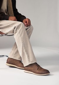 Scarpe in pelle marrone dalla texture liscia, con design allacciato e suola esterna color crema a contrasto. Indossate con pantaloni in velluto a coste beige chiaro.