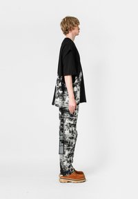 Un joven con cabello rubio corto está de lado, vistiendo pantalones de patrón blanco y negro, una camisa oversized en blanco y negro, y botas marrones.