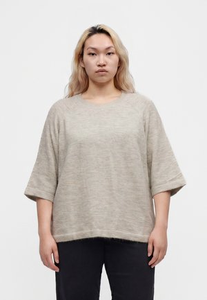 Ung person med lys blondt hår iført en løs beige sweater med halværmer og sorte bukser, stående mod en hvid baggrund.