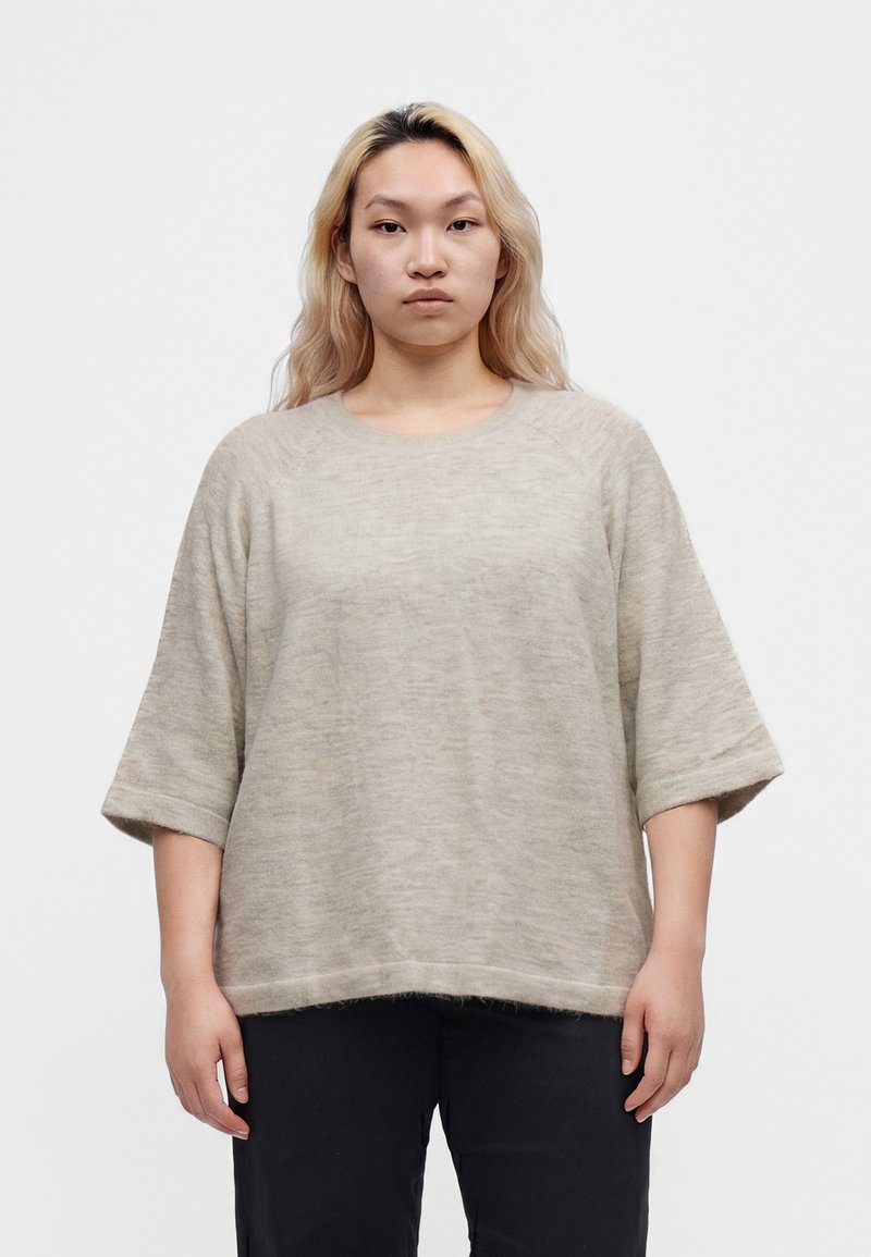 Jeune personne aux cheveux blonds clairs portant un pull beige ample à manches mi-longues et un pantalon noir, debout devant un fond blanc.