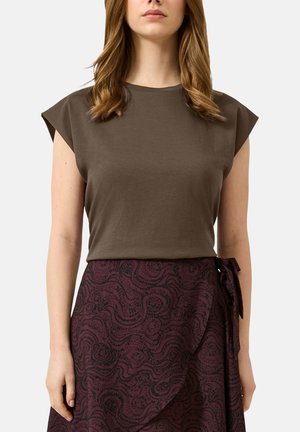 Femme portant un haut marron sans manches et une jupe portefeuille violet foncé à motifs, nouée à la taille, debout devant un fond uni.