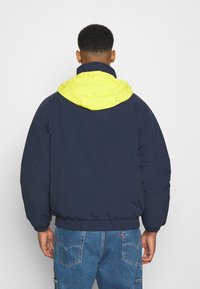 Marineblauwe donzen jas met een felgele capuchon, met een comfortabele pasvorm, elastische manchetten en tailleband, gemaakt van glad, lichtgewicht materiaal.