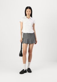 Chemise blanche à manches courtes avec un col, jupe plissée grise, mocassins noirs et un petit sac à main noir ; le mannequin se tient devant un fond blanc.