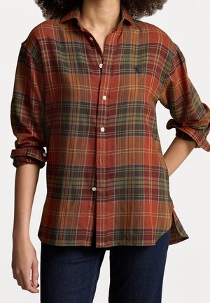 Femme portant une chemise à manches longues à carreaux orange et verte, boutonnée, aux manches retroussées, et un jean bleu foncé, sur fond blanc.