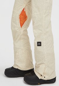 Beige sneeuwbroeken met een abstract patroon, voorzien van gaasventilatieopeningen en een logopatch op de dij. Zwarte sneeuwlaarzen worden eronder gedragen.
