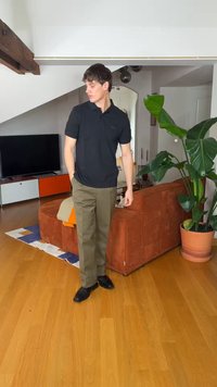 Άνδρας με μαύρο πόλο και ελαιδί παντελόνι στέκεται σε μοντέρνο σαλόνι με ξύλινο πάτωμα, καφέ-κόκκινο καναπέ, μεγάλο πράσινο φυτό και μονάδα τηλεόρασης.