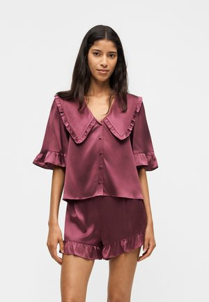 Femme portant un haut boutonné mauve brillant avec un large col à volants et un short assorti aux ourlets à volants, debout devant un fond uni.