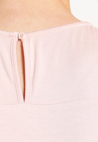 Top rosa claro con escote redondo y un corte en forma de llave en la parte posterior, terminado con un cierre de botón simple. Tejido suave y texturizado.