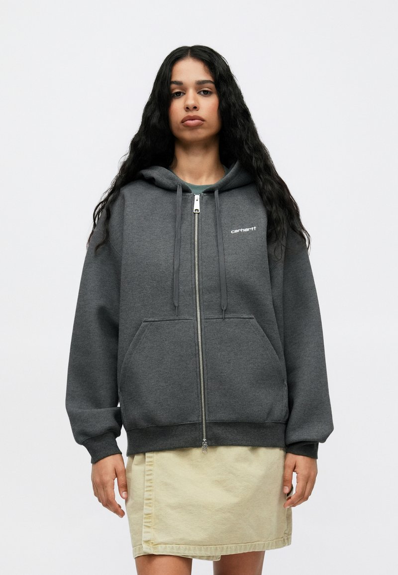 Grå zip-up hoodie lavet af blød stof, der har en stor hætte, forlommer og hvidt logo på venstre bryst. Bæres med en lys beige nederdel.