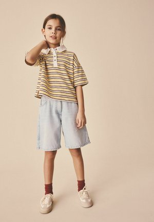 Barn iført en gulstribet polo, lyse denimshorts, røde sokker og beige sneakers står op ad en beige baggrund.