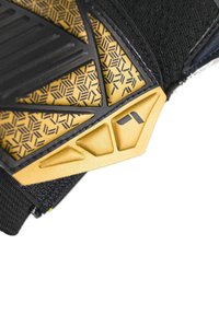 Primo piano di un guanto sportivo nero e oro con motivi geometrici testurizzati e un logo parziale sulla fascia del polso.