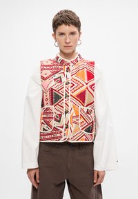 Többszínű mellény, amely bonyolult geometriai mintákat tartalmaz piros, rózsaszín és fekete színben, krém háttéren, magas gallérral és elől gombokkal.