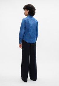 Camisa de jeans azul, com mangas longas, punhos abotoados e bainha arredondada. Combinada com calças pretas de cintura alta e perna larga.