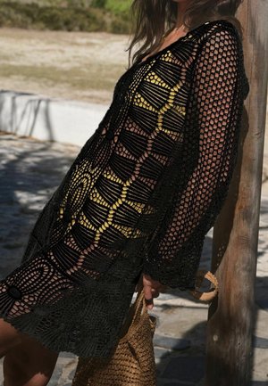 Robe noire en crochet à manches longues, présentant un motif ajouré et des accents dorés. Le mannequin tient un sac en paille tressée, debout contre un poteau en bois.