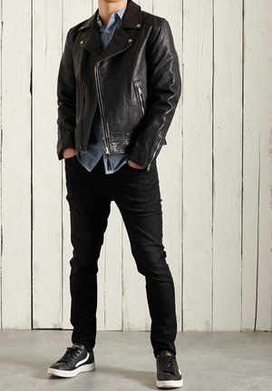Chaqueta de motociclista de cuero negro con herrajes plateados, superpuesta a una camisa de rayas azules y blancas. Combinada con jeans negros y zapatillas negras.