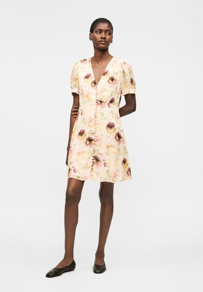 Γυναίκα με κοντομάνικο, φλοράλ φόρεμα με V-λαιμόκοψη, κουμπιά μπροστά και μαύρα φλατ παπούτσια, στέκεται μπροστά σε απλό λευκό φόντο.