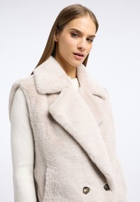 Kamizelka z futra syntetycznego w jasnym beżu z szerokim kołnierzem i podwójnym zapięciem na guziki. Miękka tekstura, bezrękawnik i subtelny połysk.