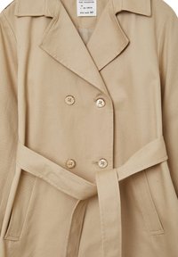 Beige Doppelreiher mit Reverskragen, ausgestattet mit zwei Knöpfen, einem Bindegürtel und Seitentaschen. Strukturierter Stoff mit glatter Oberfläche.