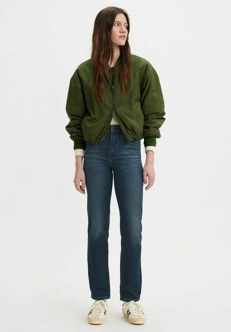 Olivgrön bomberjacka, dragkedjestängning, ribbade ärmar, i kombination med mörkblå jeans och ljusa sneakers. Enkel design, avslappnad stil.
