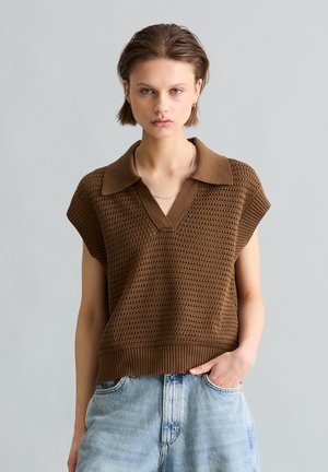 Młoda osoba z krótkimi włosami, ubrana w brązowy teksturowany bezrękawnik z kołnierzykiem i jasnoniebieskie dżinsy, stojąca na jednolitym tle.