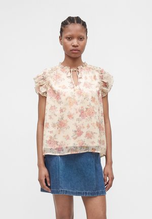 Kvinde med fletninger iført en gennemsigtig blomsterbluse med flæser på ærmerne og bindebånd ved halsen, kombineret med et blåt denimskørt.