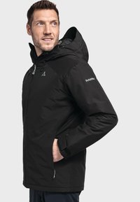 Zwarte jas met een capuchon, uitgerust met een rits aan de voorkant, twee zijzakken en een logo op de mouw. Materiaal lijkt glad met een matte afwerking.