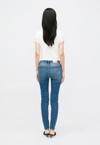 Γυναίκα με μακριά μαύρα μαλλιά, φορώντας λευκή κοντομάνικη μπλούζα, μπλε στενές τζιν παντελόνες με πίσω τσέπες και μαύρες ψηλοτάκουνες σαγιονάρες, που στέκεται σε λευκό φόντο.