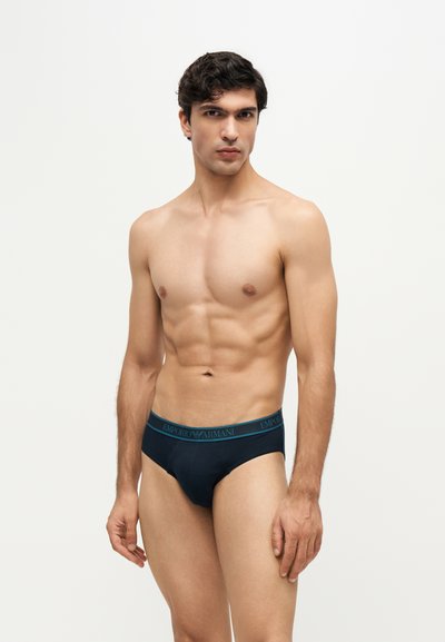 Jeune homme en forme aux cheveux foncés portant des slips noirs Emporio Armani sur un fond clair uni.