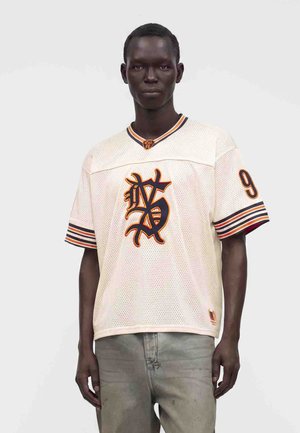 Sort mand iført en hvid net-sportsjersey med orange og sort logo og nummer ni på ærmet, kombineret med grå bukser.