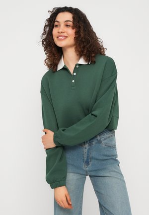 Donna con capelli ricci che indossa una maglia verde a maniche lunghe con colletto a polo e jeans azzurri chiari, in piedi con le braccia incrociate davanti.