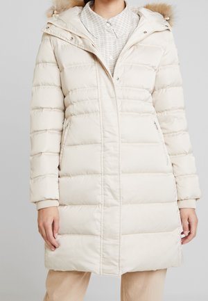 Femme portant un manteau matelassé mi-longue blanc cassé avec poches zippées et une capuche bordée de fourrure sur une chemise légère à motifs.