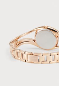 Hodinky Swarovski v růžovém zlatu s kovovým náramkem, hladkým kulatým pouzdrem a minimalistickým designem. Na sponě je vyryto logo.