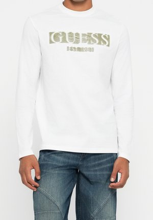 Mężczyzna w białej koszuli z długim rękawem z logo "GUESS" w złotym brokacie i niebieskich dżinsach na jednolitym tle.