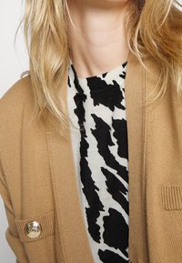 Beige knit cardigan with a round, gold logo button, layered over a white shirt with a black abstract animal print pattern.