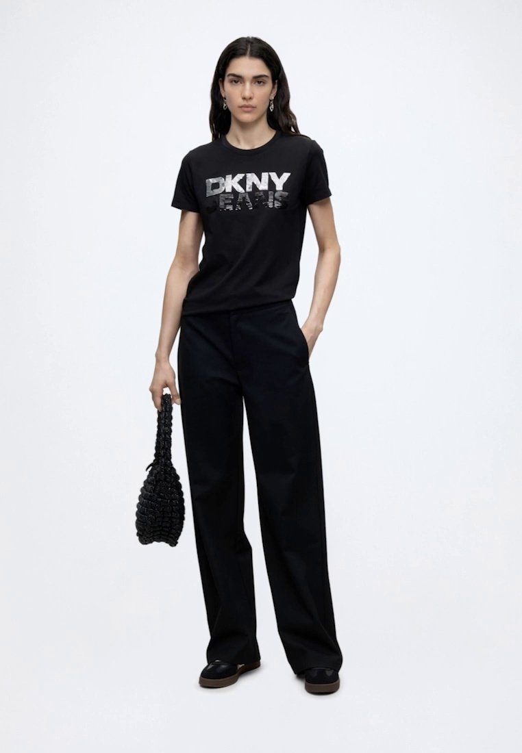 Жена, облечена в черна тениска DKNY и широки черни панталони, държаща черна плетена чанта, стои пред обикновен бял фон.