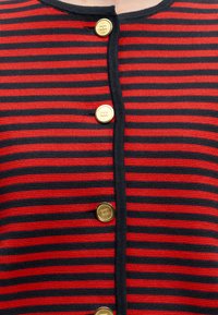 Pull en maille à rayures horizontales rouges et noires avec des boutons ronds dorés sur le devant et un bord noir le long de la ligne de boutons.