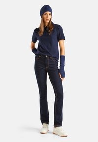 Camisa polo azul marino, jeans oscuros con costuras naranjas, gorro de punto azul, guantes sin dedos y zapatillas blancas con suelas de goma.