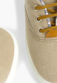 Baskets en toile beige avec une finition texturée, bout rond, semelle en caoutchouc blanc et lacets marron contrastants passés dans des œillets en métal.