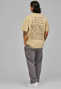 Tricou oversized bej cu un text grafic mare de culoare maro închis pe spate. Purtați-l cu pantaloni gri și adidași albi.