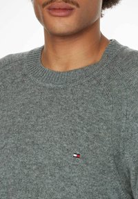 Grauer Strickpullover mit einem gerippten runden Halsausschnitt und Raglanärmeln. Verfügt über ein kleines rot-, weiß- und blaues Logo auf der linken Brust.