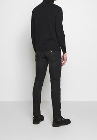 Svart polotröja med långa ärmar, kombinerad med smala svarta jeans och ankle boots, sedd bakifrån mot en ljus bakgrund.