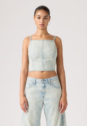 Žena oblečená v svetlomodrom rifľovom krátkom topánku a vysokých džínsach, stojaca pred jednoduchým svetlým pozadím.