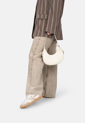 Witte leren handtas met een gebogen vorm en zilverkleurige hardware, vastgehouden door een persoon in een gestreepte bruine blazer en beige wijde broek.