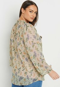 Blusa floreale trasparente in colori pastello, con maniche lunghe e gonfie, scollatura a volant e vari motivi floreali su uno sfondo puntinato.